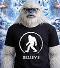 Furry white yeti wearing glasses and a black t-shirt with a "Believe" Bigfoot logo.