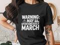 Black t-shirt with white text "WARNING: MAY SPONTANEOUSLY MARCH" and boot icon.
