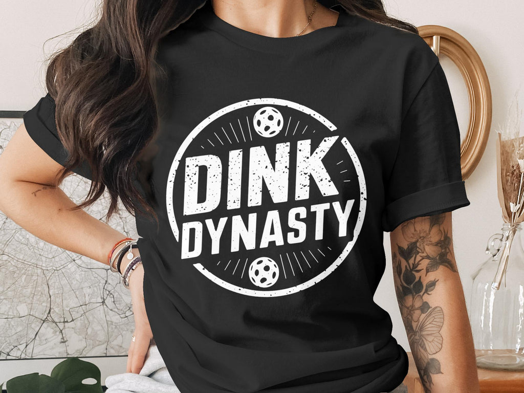 White "DINK DYNASTY" text with soccer balls on a black t-shirt.