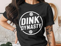 White "DINK DYNASTY" text with soccer balls on a black t-shirt.