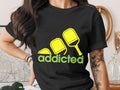 Black t-shirt with "addicted" text and pickleball paddles graphic.