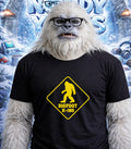 Yeti wearing glasses and a "Bigfoot X-ing" t-shirt against a frosty background.